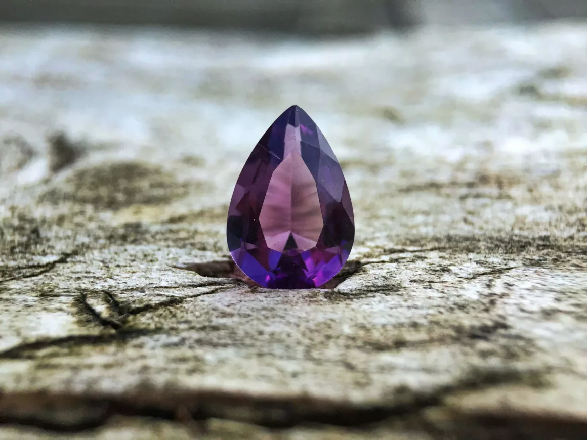 A close-up image from Gemstones Insider showcases a pear-shaped, faceted purple gemstone—possibly an amethyst—resting on a textured wooden surface. The photo features sharp detail in the foreground with a softly blurred background, highlighting the gem’s exquisite color and cut.