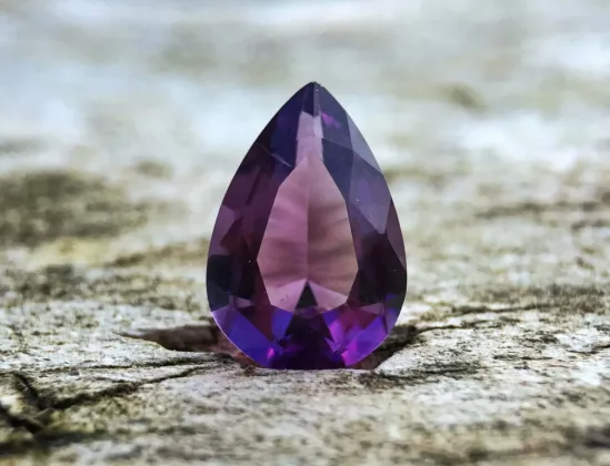 A close-up image from Gemstones Insider showcases a pear-shaped, faceted purple gemstone—possibly an amethyst—resting on a textured wooden surface. The photo features sharp detail in the foreground with a softly blurred background, highlighting the gem’s exquisite color and cut.