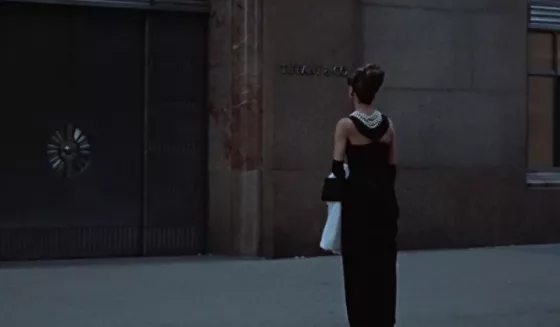 Audrey Hepburn in a black dress with a multi-strand pearl necklace, elegant updo, and black gloves, standing outside a jewelry store window in a classic scene from Breakfast at Tiffany’s, showcasing pearls on screen.