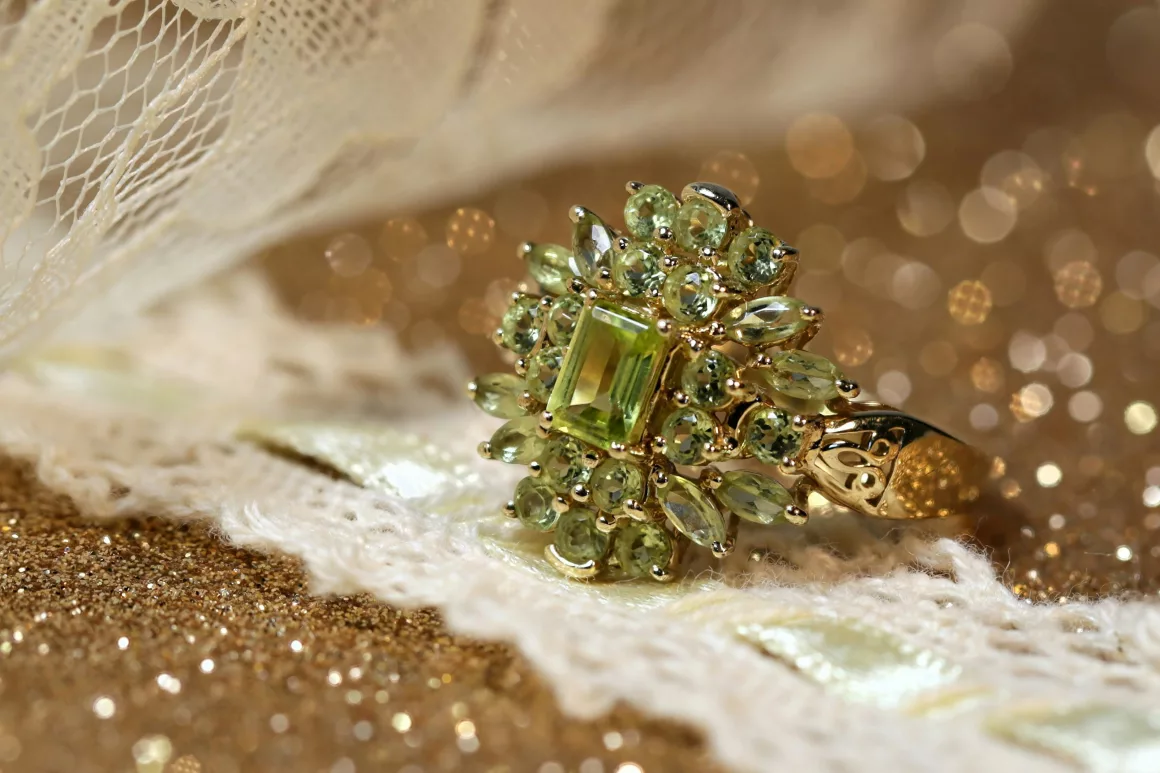 A vintage-style gold ring set with a rectangular peridot gemstone surrounded by smaller marquise and round peridot stones, displayed on a lace and glittering gold background.