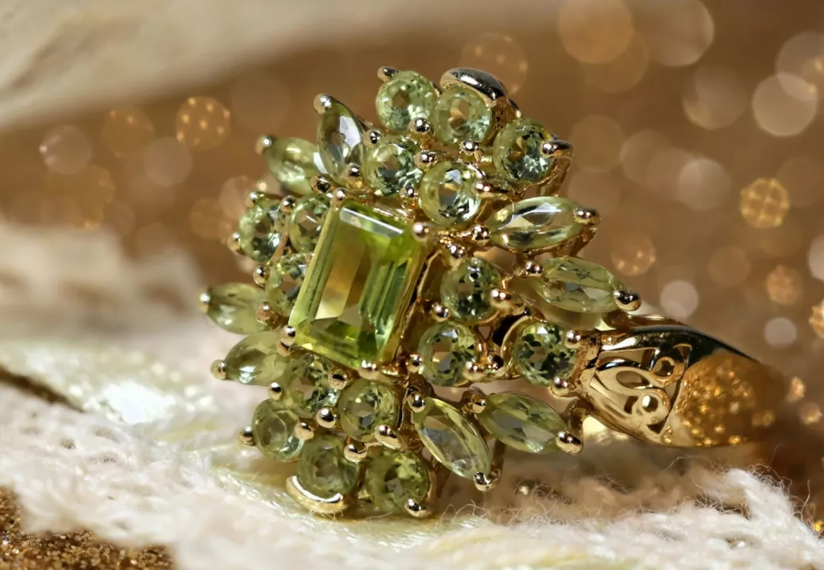 A vintage-style gold ring set with a rectangular peridot gemstone surrounded by smaller marquise and round peridot stones, displayed on a lace and glittering gold background.