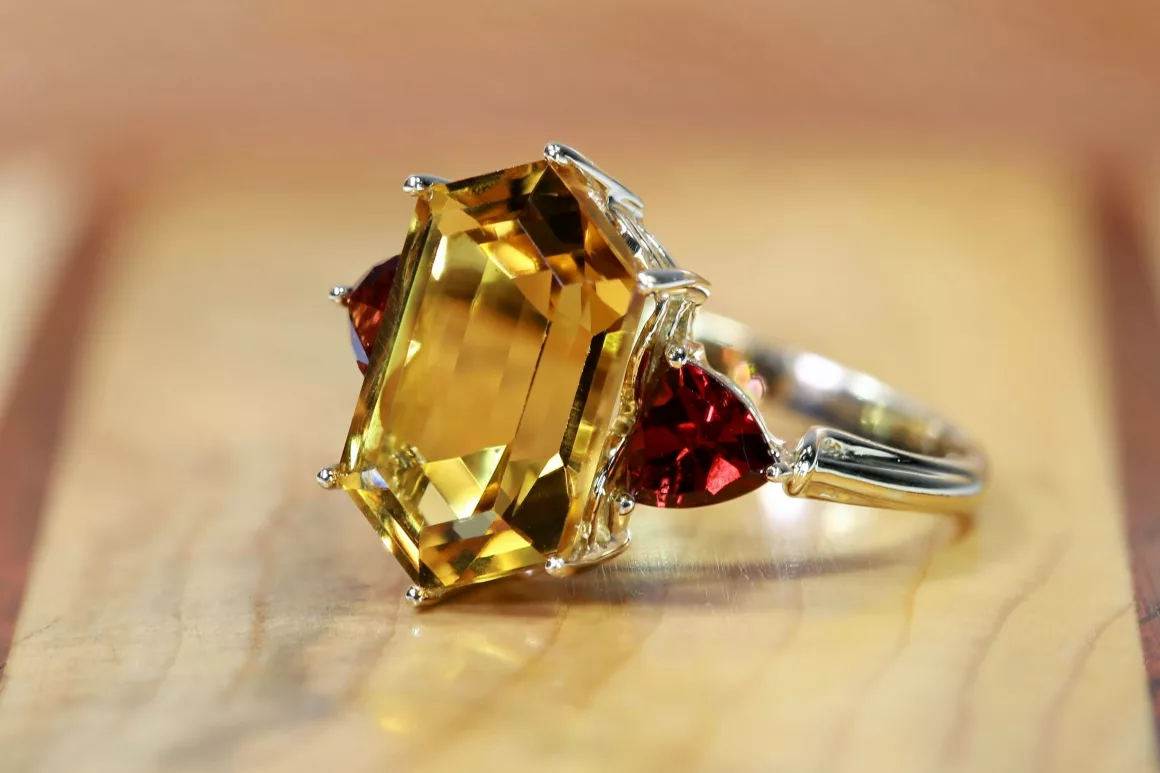 A close-up of a gemstone engagement ring featuring a large rectangular yellow gemstone flanked by two deep red stones, illustrating 2025 jewellery trends.