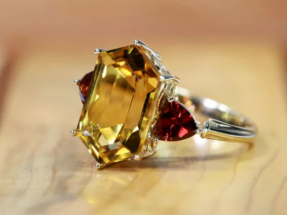 A close-up of a gemstone engagement ring featuring a large rectangular yellow gemstone flanked by two deep red stones, illustrating 2025 jewellery trends.