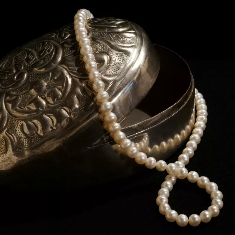 A classic strand of white pearls elegantly draped over an ornate, engraved silver jewelry box against a black background, symbolizing june birthstones and pearls.