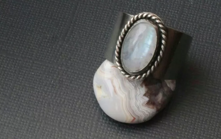 A wide silver ring featuring an oval moonstone with a braided bezel setting, elegantly displayed on a banded agate stone against a textured dark background.