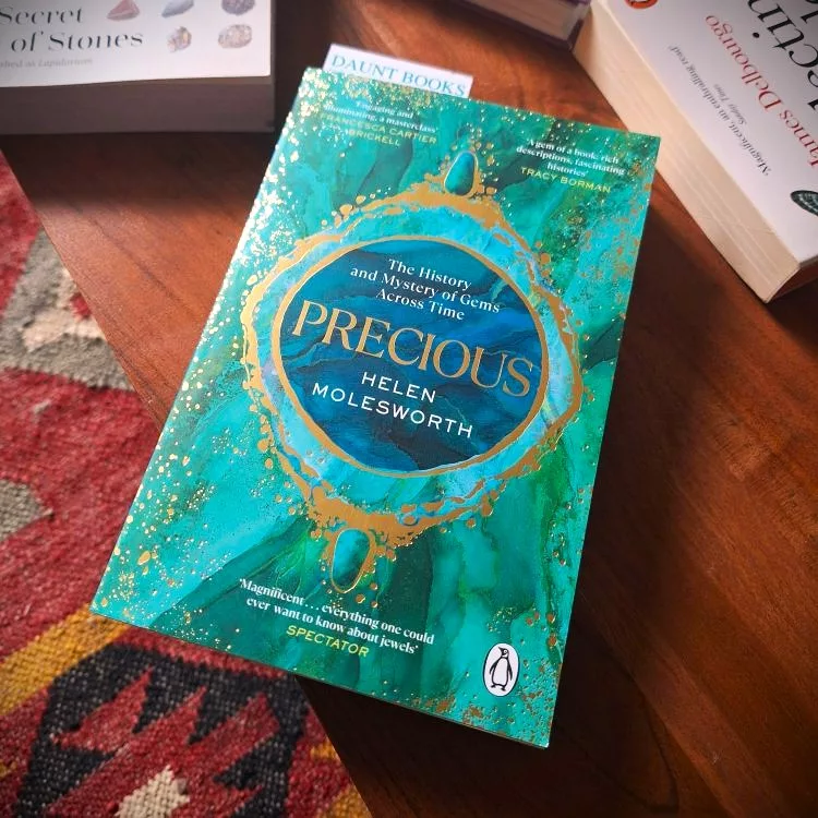 Cover of the book Precious: The History and Mystery of Gems Across Time by Helen Molesworth, displayed on a wooden table with other gemstone books nearby.