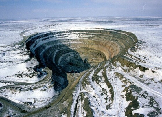A vast open-pit diamond mine in a snowy landscape, with terraced circular walls and winding access roads, illustrating the large-scale extraction operations central to the impact of sanctions and the gemstone market.