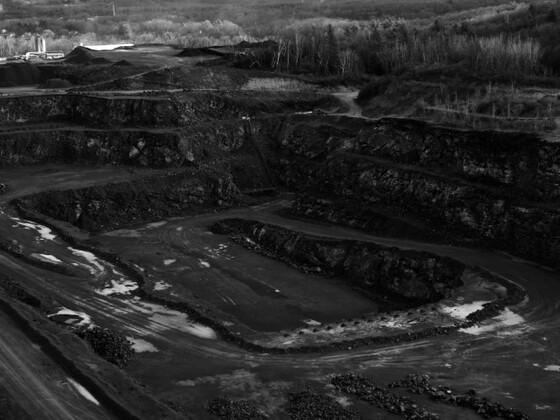 A wide, terraced open-pit mine surrounded by forest, with exposed earth and rock layers, illustrating the environmental and social complexities tied to the politics of gemstone mining.