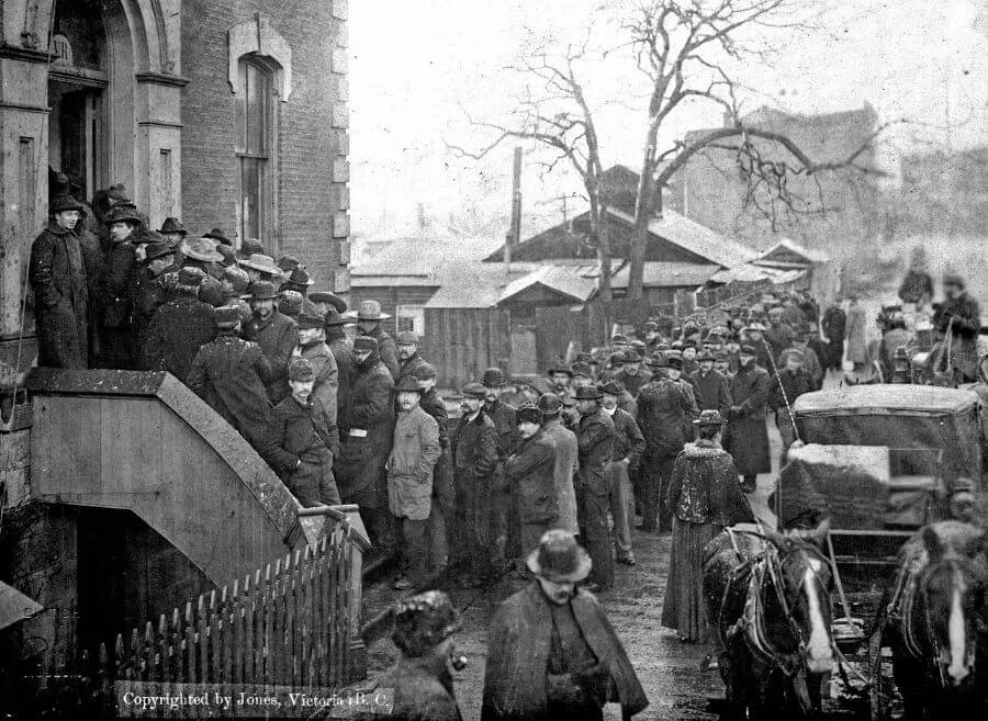 A long line of men in heavy coats and hats standing outside a building, waiting to buy miner’s licenses during the Klondike Gold Rush, with horse-drawn carriages and wooden shacks visible in the background on a cold, overcast day.