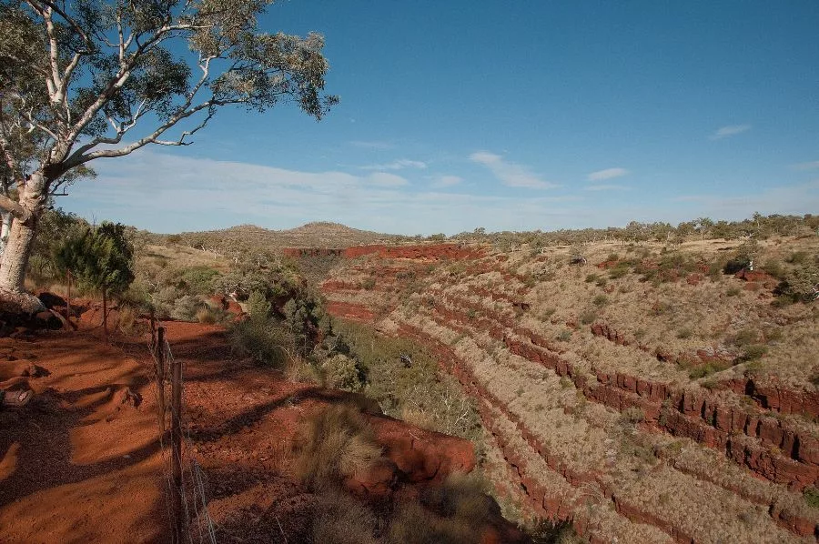 A panoramic view of Dales Gorge in Karijini National Park, Western Australia, featuring dramatic red rock layers, sparse vegetation, and a clear blue sky—an iconic example of Geological and Mineral Destinations in Australia.