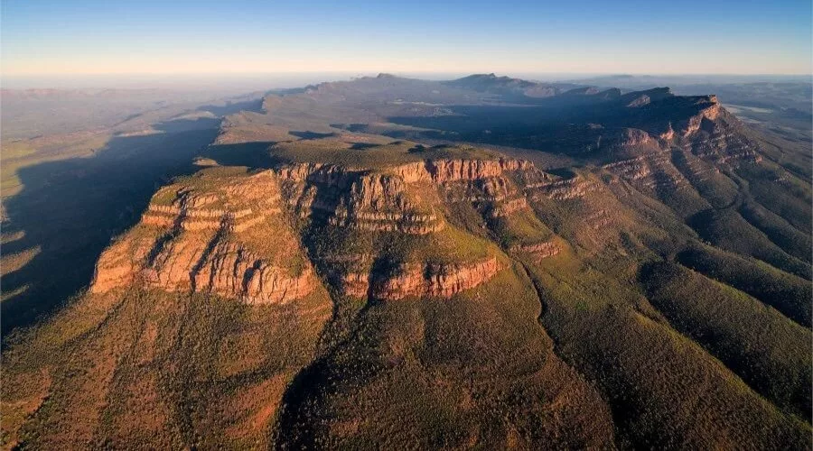 A panoramic aerial view of the rugged, layered red rock formations and expansive valleys of the Flinders Ranges in South Australia, bathed in warm sunlight and stretching toward distant hills under a clear blue sky—an iconic example of Geological and Mineral Destinations in Australia.