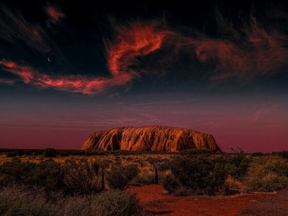 Uluru (Ayers Rock) glowing red at sunset beneath dramatic crimson clouds and a crescent moon, surrounded by desert vegetation, exemplifying iconic Geological and Mineral Destinations in Australia.