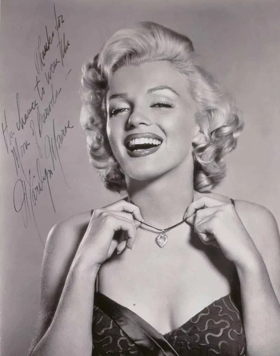 A classic black-and-white portrait of Marilyn Monroe wearing an elegant strapless dress, holding up a necklace featuring the famous Moon of Baroda Diamond, showcasing the glamour and allure of Marilyn Monroe's jewellery collection.