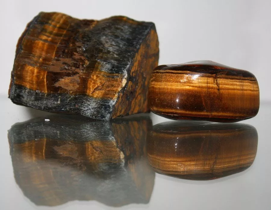 Two pieces of Tiger’s Eye, one raw and one polished, displaying golden-brown chatoyancy and fibrous bands with reflective surface detail.