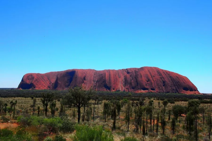 Uluru (Ayers Rock) glowing red at sunset beneath dramatic crimson clouds and a crescent moon, surrounded by desert vegetation, exemplifying iconic Geological and Mineral Destinations in Australia.