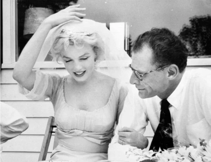 Marilyn Monroe smiles in a soft wedding dress and veil, seated beside Arthur Miller, who looks at her fondly while wearing his gold wedding band—part of Marilyn Monroe’s jewellery collection legacy.