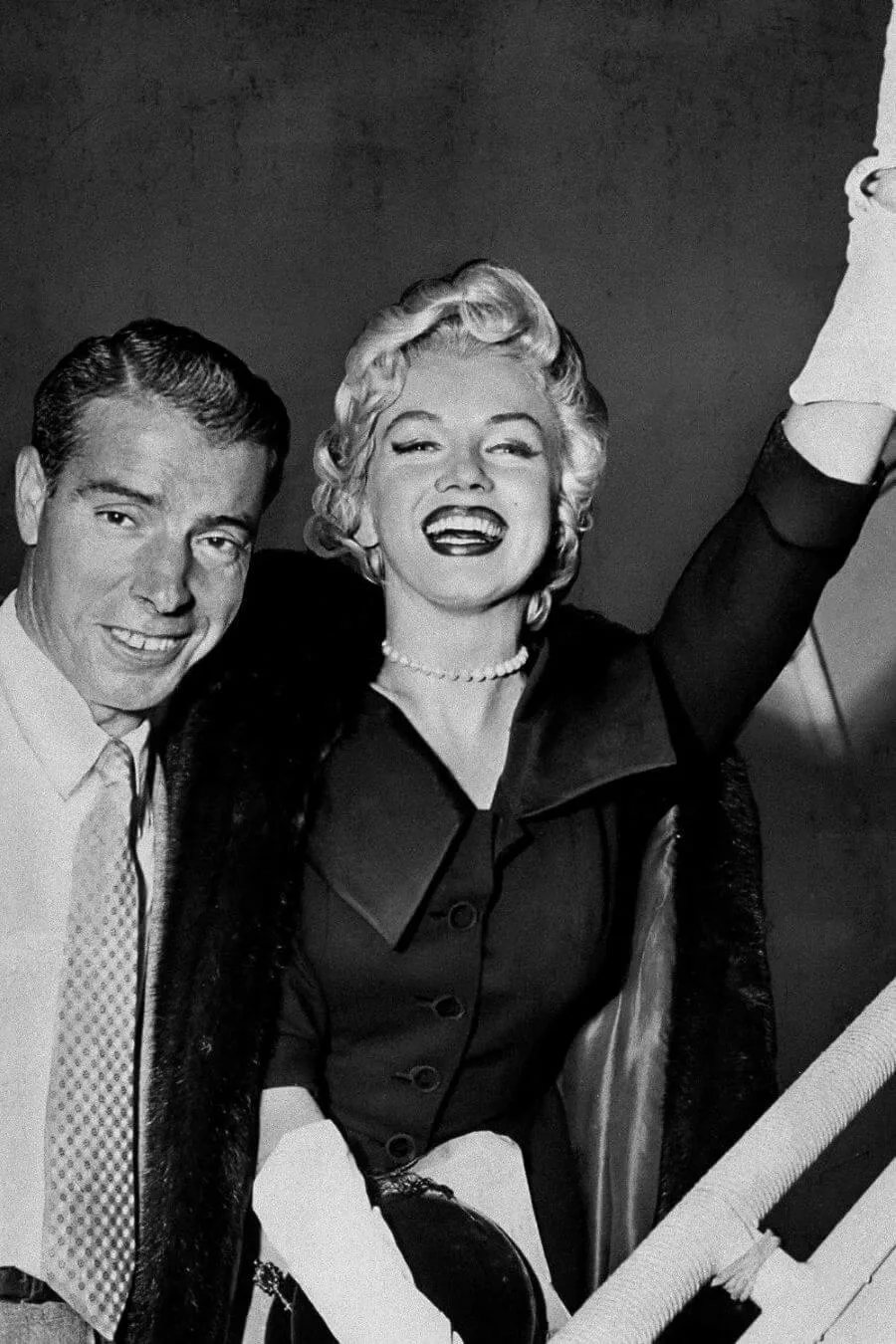 Marilyn Monroe smiles and waves while wearing a dark dress, white gloves, and the Mikimoto Pearl Necklace, standing beside Joe DiMaggio in a black-and-white photograph.