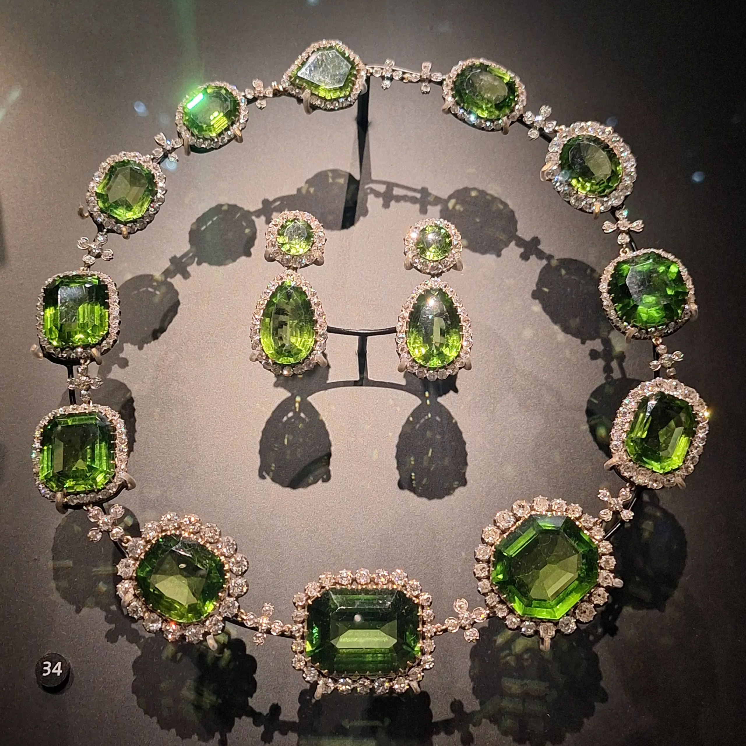 A luxurious set of green peridot jewelry including a necklace and earrings, displayed under lighting at the V&A Museum.