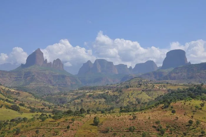 Expansive view of the rugged Simien Mountains with steep cliffs, ridges, and scattered vegetation under a blue sky with clouds.