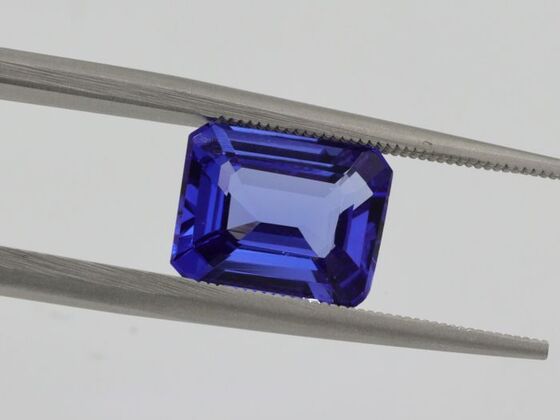 A vivid emerald-cut blue gemstone held by tweezers, illustrating quality and clarity factors that influence Gemstone Prices.